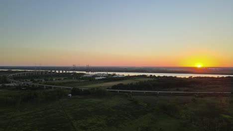 dolly right flying over green fields and zarate brazo largo road and railway complex cable-stayed bridge crossing parana river at sunset, entre rios, argentina