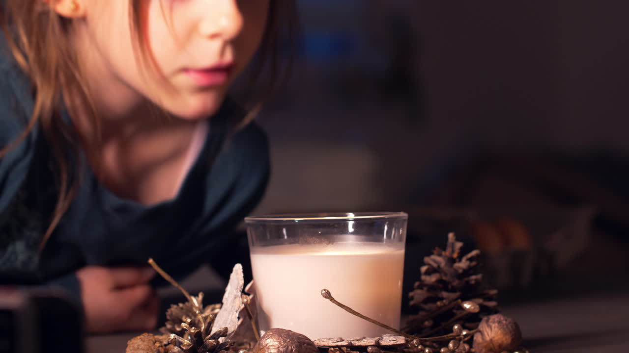 A young girl blows into a decorative candle on the table in a glass jar, catches the smoke of the candle with her hand, static