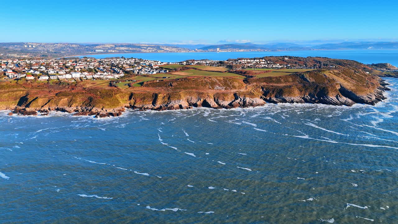 A cena mostra uma área costeira com várias casas situadas em uma encosta acima da água. Ondas quebram contra a costa rochosa, e a água se estende até o horizonte.