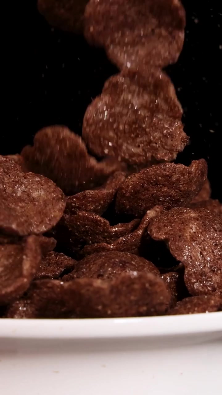 Slow motion vertical shot of chocolate cereal flakes cascading into a white ceramic bowl.