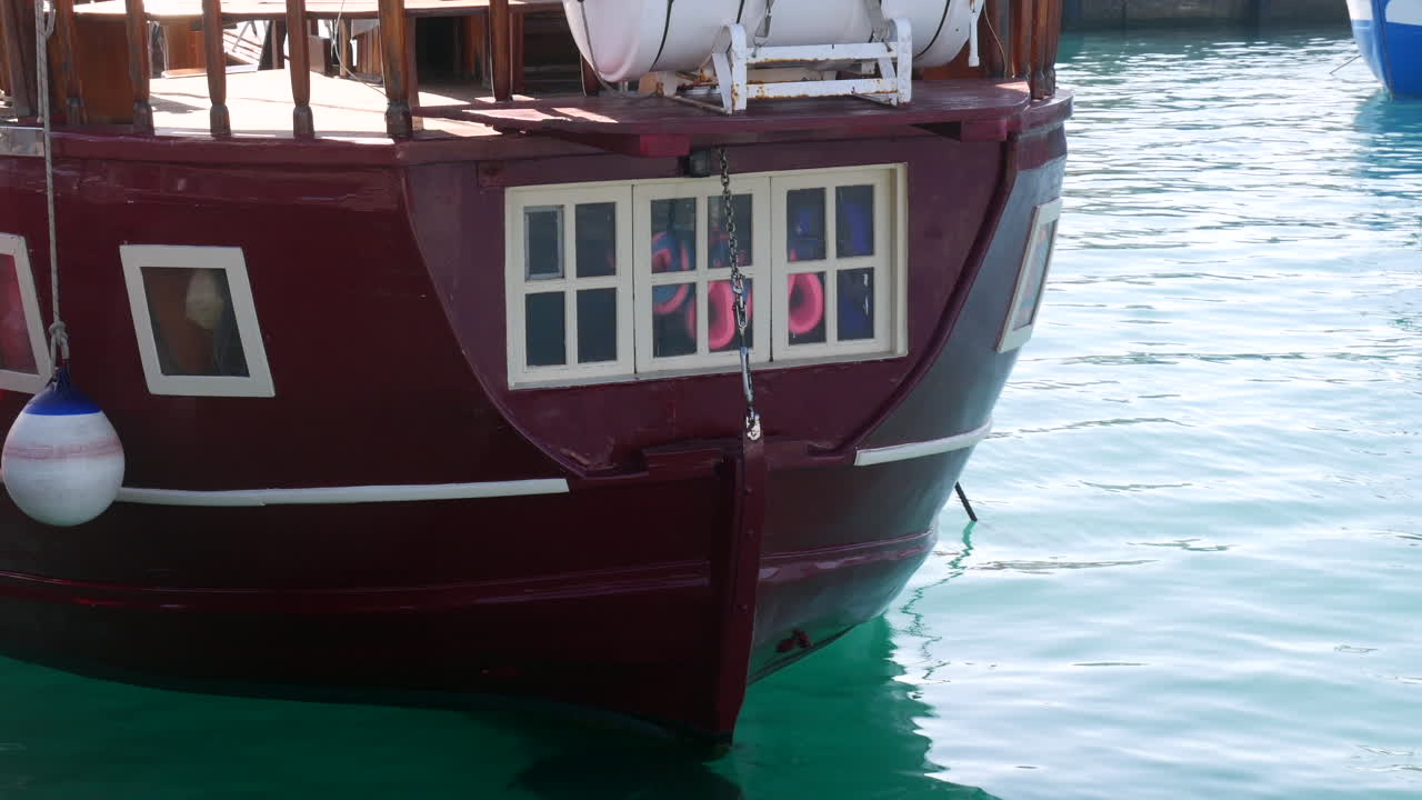 Close-up of a sail boat stern with quaint windows reflecting life aboard, anchored in tranquil turquoise waters, hinting at maritime pirate adventures