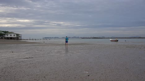 Escena-De-Un-Hombre-Caminando-Sobre-La-Arena-De-Una-Playa-De-Marea-Baja-Con-El-Telón-De-Fondo-De-Una-Casa,-Un-Barco-Y-Una-Serena-Vista-Al-Mar