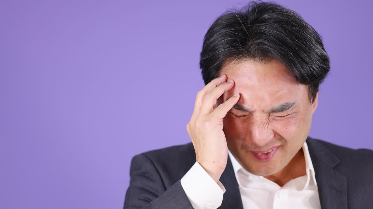 Asian businessman in suit massages temples, eyes closed, under bright studio lighting, purple backdrop