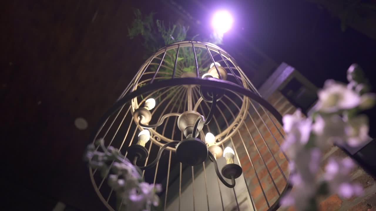 A low-angle close-up of a vintage cage chandelier or lantern hanging above white flowers and a brick wall, creating a romantic night ambiance