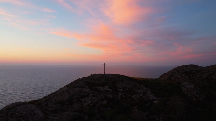 Una croce solitaria si erge su una collina, stagliata contro un'alba vibrante sull'oceano. Ideale per temi di riflessione o spiritualità.
