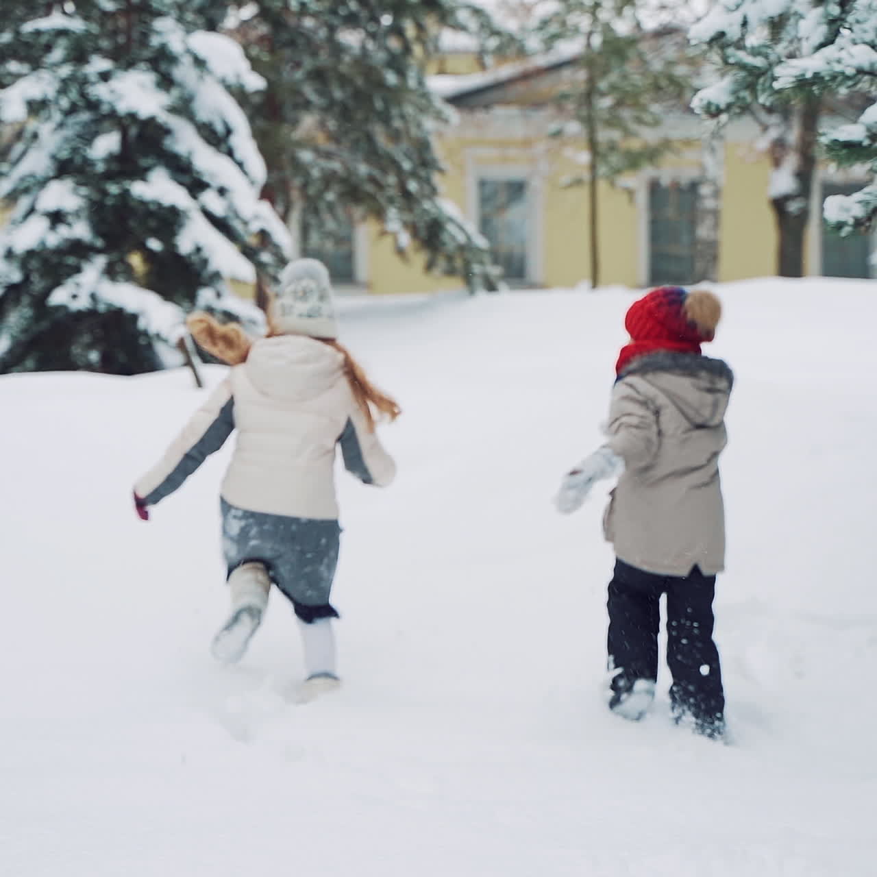 Two active little girls are running and playing in the white snow outdoors. Children in warm clothes relax together on the snowy background with a small yellow building.