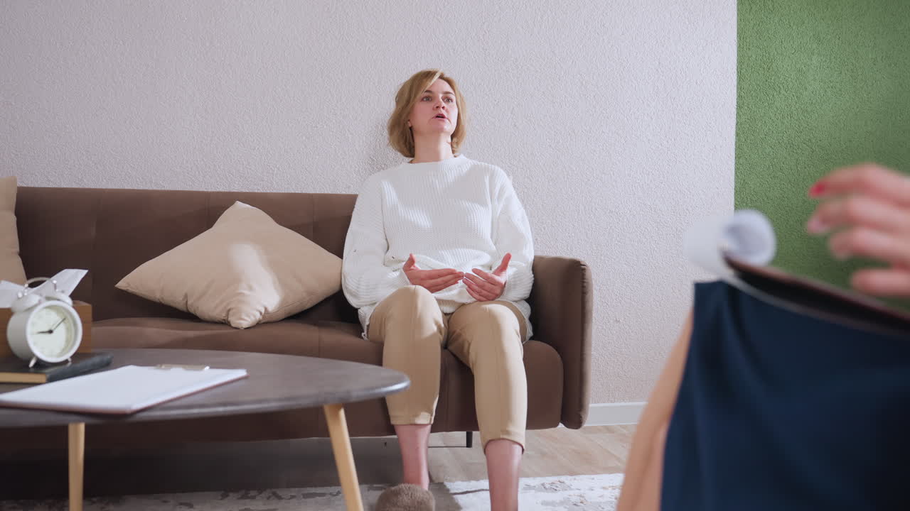 Zoomed out view of participant speaking to therapist taking notes, cozy counseling room with green chair, brown sofa, table with clock and pen pack, slippers and pillow details visible