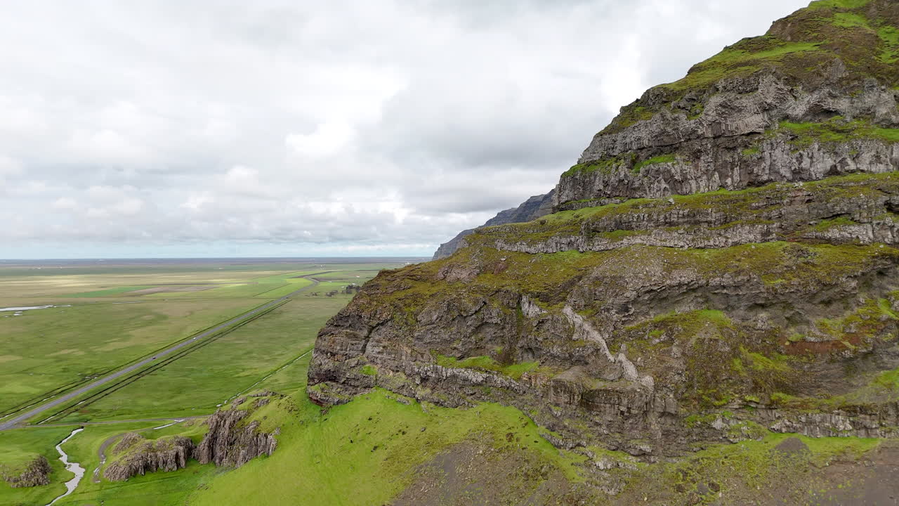 Aerial view of vibrant green fields stretching below steep moss-covered cliffs in Suðurland, Iceland, with winding streams and dramatic shadows shaping the vast volcanic landscape