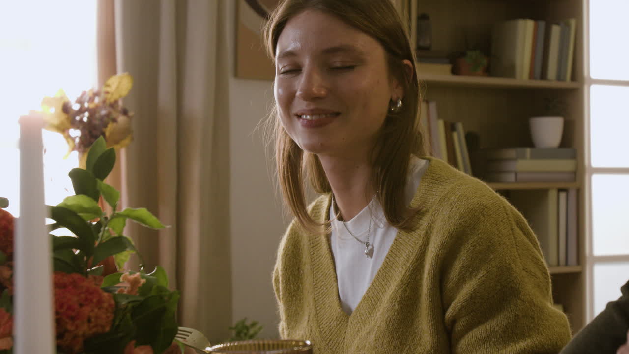 A woman sits at a table with a candle and bookshelf in the background