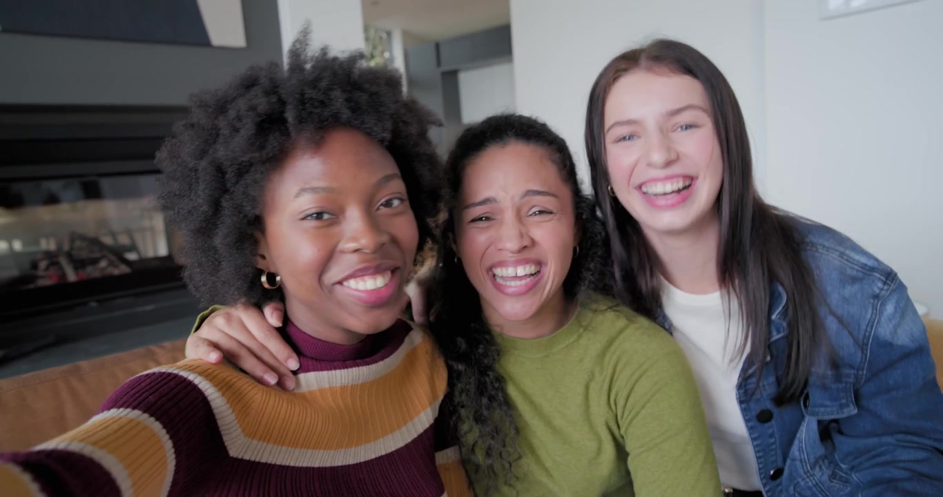Tre amiche allungano il telefono, posano, scattano un selfie e ridono sul divano di casa.