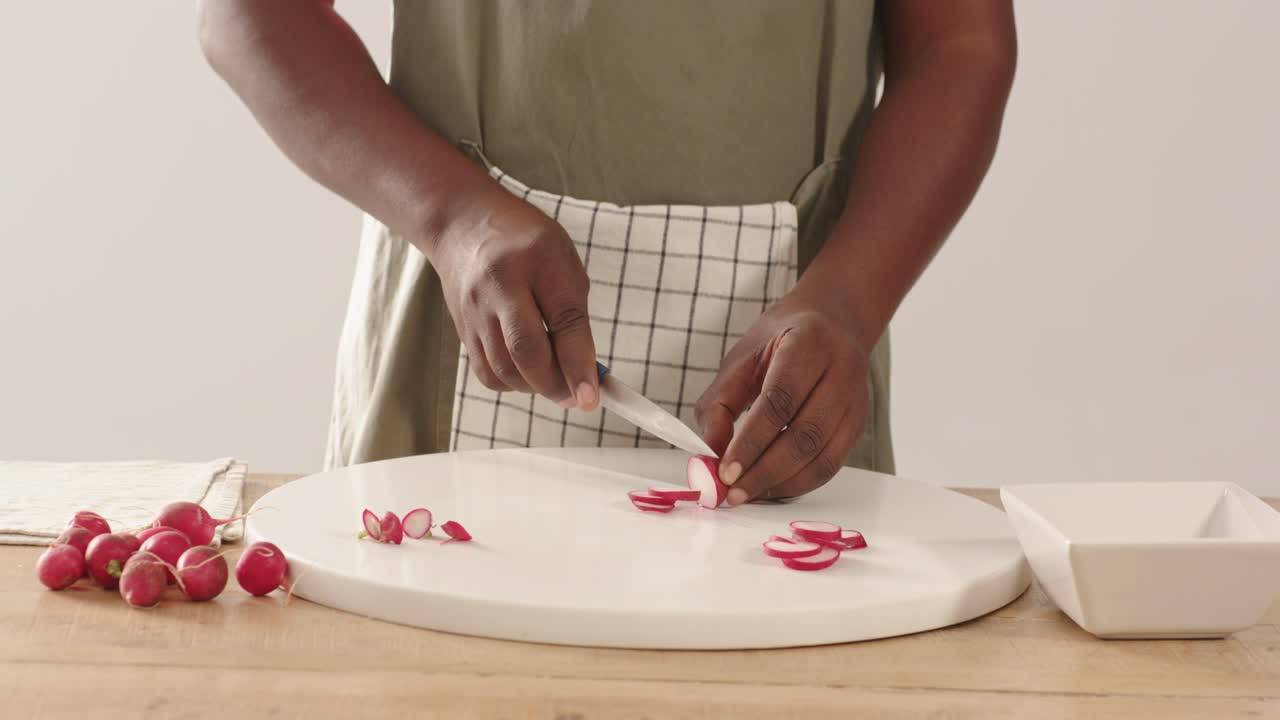 Uomo afroamericano che prende un ravanello, lo stabilizza e lo affetta con un coltello sul bancone per preparare un piatto, indossando un grembiule.