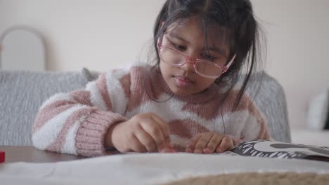 a young girl in glasses is coloring in a book at a table.