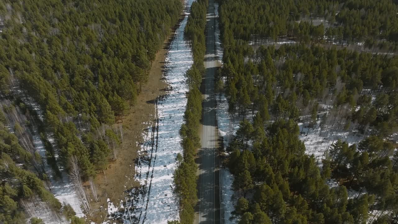 Droneoptagelser af en lige asfaltvej, der skærer gennem taigaskov med pletter af smeltende sne i det tidlige forår, Buryatia, Sibirien, Rusland, set oppefra.
