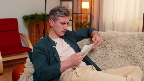 Young man reacts with stress and worry after opening envelope with credit bill at home indoors
