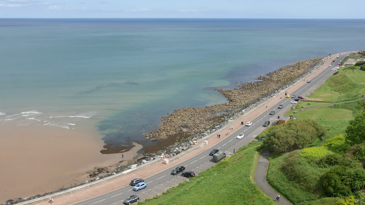 A road runs along the rocky coastline in Scarborough, North Yorkshire, England, with cars driving and people walking beside the calm sea and green hillside