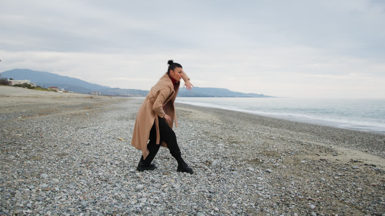 On the Shoreline in Winter a Woman Dances to Classical Music Wearing Brown Coat