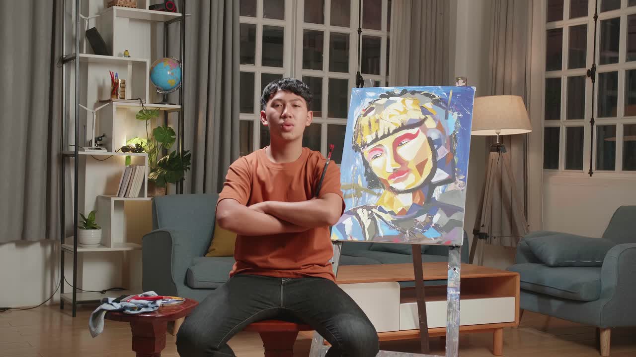 Asian Artist Boy Sitting On Round Wooden Chair Without Backrest Holding Paintbrush Mixed Colour Wipe The Sweat Before Crossing His Arms And Smiling After Finish Painting A Girl On The Canvas