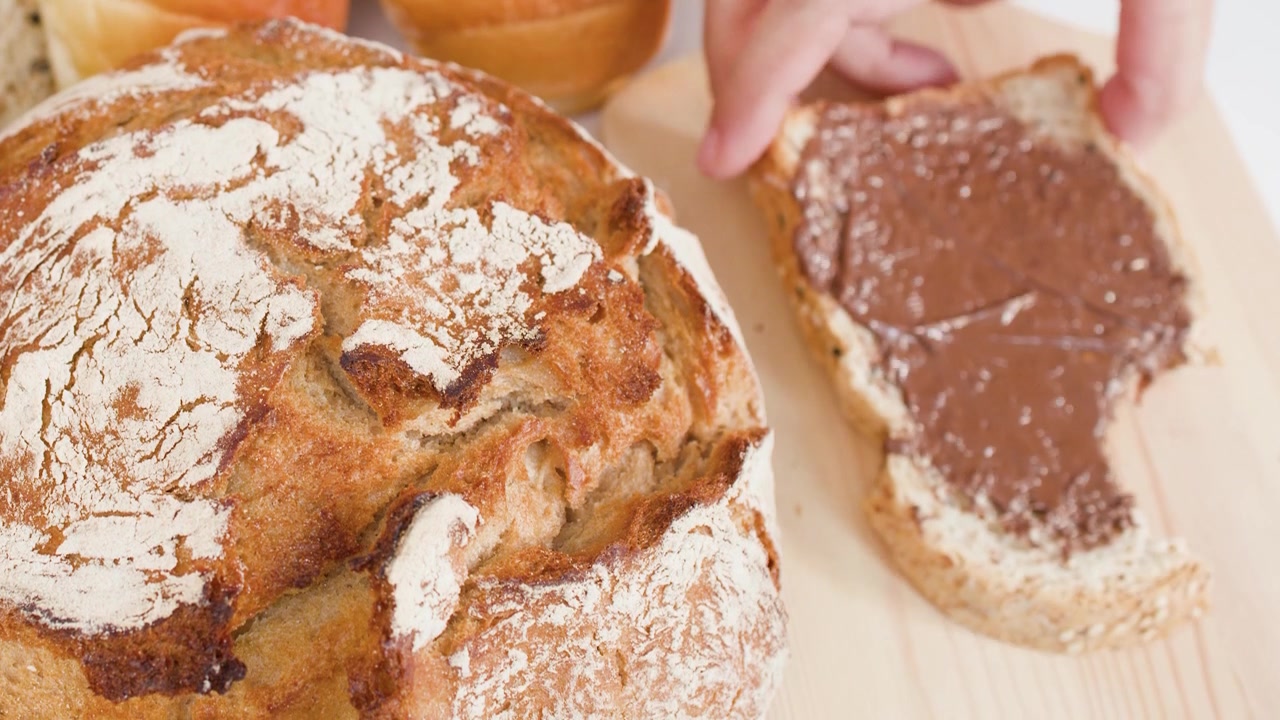 Close-up of hand lifting chocolate-covered bread slice from wooden board among sourdough and rolls.