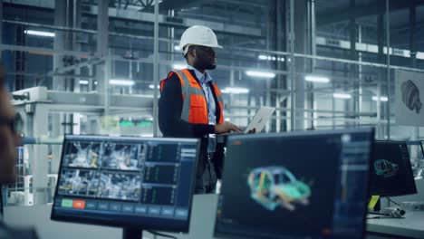 african american engineer using laptop computer and looking out of the office at a car assembly plant. vehicle factory line operator working at desk, overviewing autonomous electric car production.