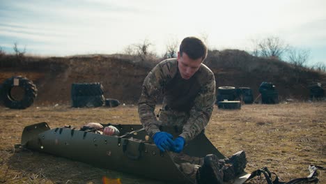 Confident-young-male-military-man-in-camouflage-and-medical-blue-gloves-straps-and-stabilizes-an-unconscious-man-on-a-special-green-army-stretcher-during-combat-exercises-at-a-training-ground-in-the-steppe