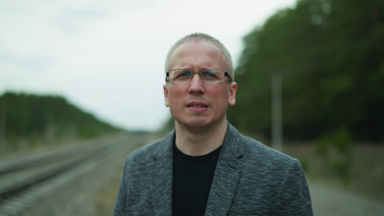 vista de primer plano de un hombre con gafas y una chaqueta gris de pie en una vía ferroviaria, mirando a la distancia con una expresión seria