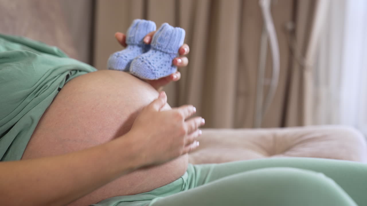 Big belly of a pregnant woman. Future mother holding little knitted socks on her stomach. Close-up. Side view.