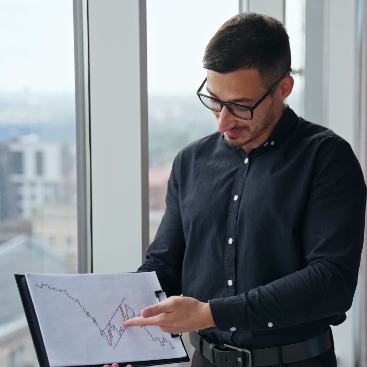 Male employee in black clothes and glasses points at documents in his hands. Caucasian man talks showing charts with a surprised face expression