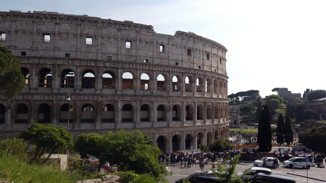 The colosseum from a different side than usual on a sunny spring day. There are few cars parked on the sides of the street. Cars are passing by. People are walking on the sidewalk.