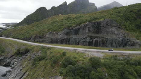 Aerial-view-showing-a-black-vehicle-moving-along-a-road-that-winds-along-the-mountains-and-the-ocean-in-Lofoten,-Norway