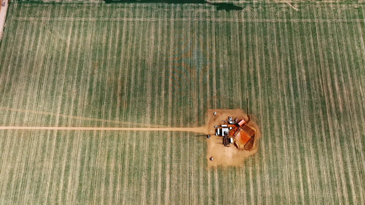 Huge field with little site for oil production in the middle. Drone footage descending above the location with drilling equipment and a square pool.