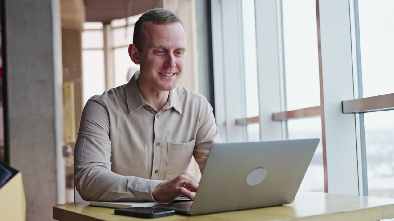 Smiling young man working at his laptop. Male employee says something and looks into window. Blurred backdrop.