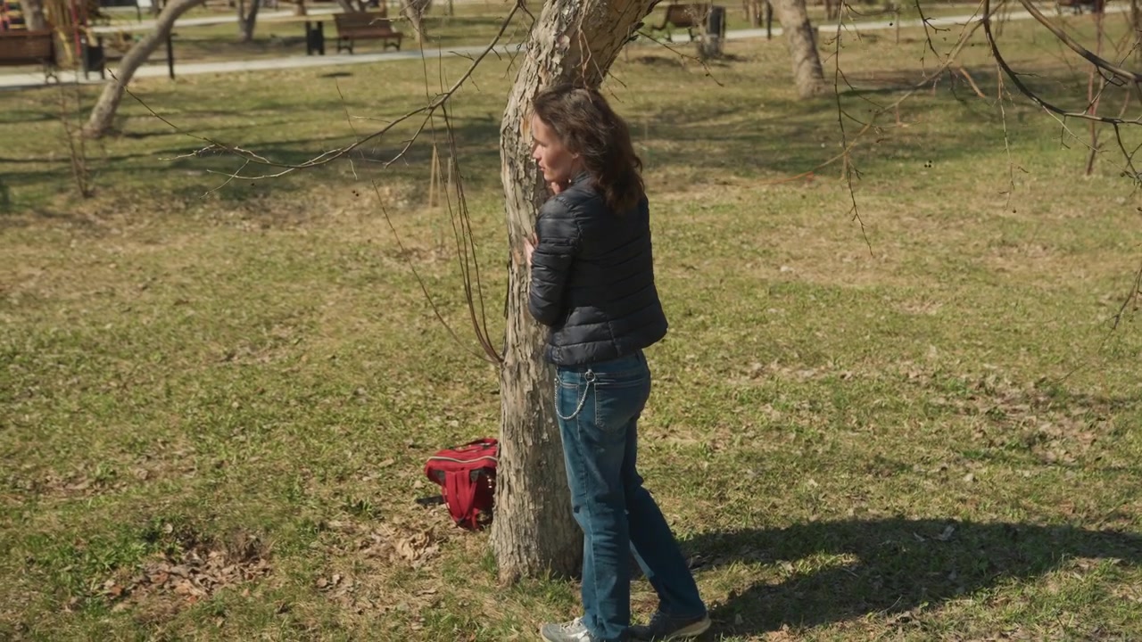 Woman gently embraces tree in outdoor park setting standing close to trunk with her hands on its bark. Woman wears casual clothing including jeans and puffer jacket while interacting softly