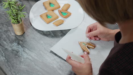 top-down view of woman decorating cookies with icing and adjusting green bead using toothpick while other cookies are on tray, scissors and decorative flower