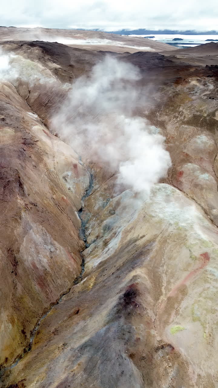 Veduta aerea con drone dei colorati pendii di riolite di Namafjall. Fumarole si innalzano da colline striate di minerali nelle tonalità del marrone, rosso e ocra. Verticale.