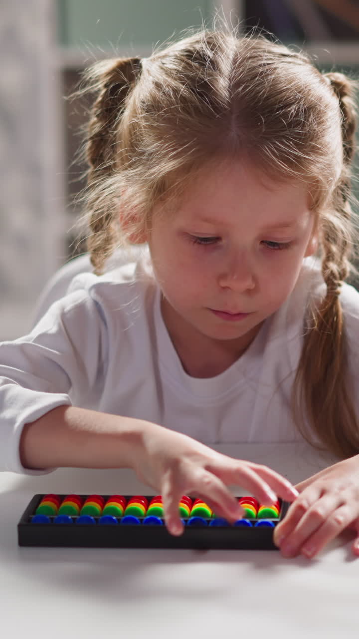 Cute little girl student with long plaits learns mental mathematics with toy abacus sitting at white desk during lesson at home closeup slow motion