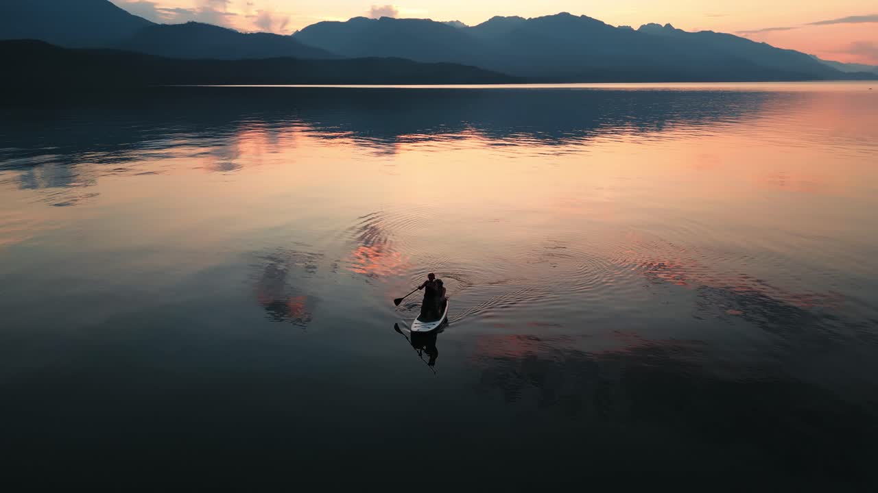 Couple sit on an inflatable paddle board as they float and paddle around the huge Harrison Lake in Chilliwack British Columbia at a golden hour sunset with orange reflections on the water