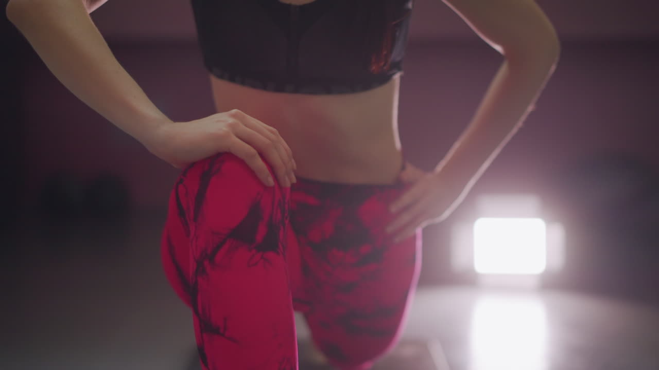 primo piano in studio di donna asiatica che allunga quadricipiti e anca, mani sui fianchi, leggings rossi fantasia e reggiseno sportivo nero, forte faretto in controluce, presenza sicura da istruttrice e posa da modella atletica, affondo