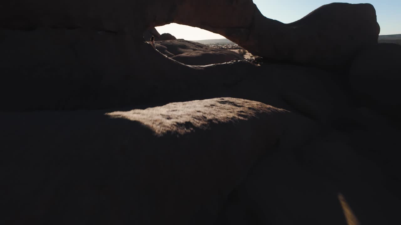 Epic FPV drone shot through the Needle gap in the rocky Namibian desert landscape of Spitzkoppe, Namibia. A lone courageous woman stands between massive stones in crisp morning light.