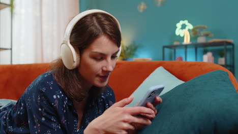 young woman in wireless headphones relaxing lying on couch at home using smartphone listening music