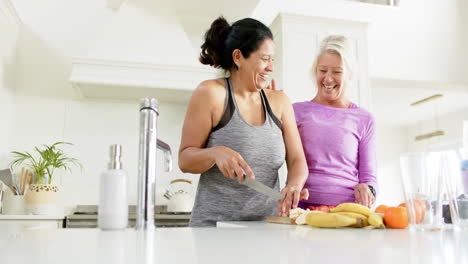 two happy diverse senior women chopping fruits and laughing in sunny kitchen, slow motion