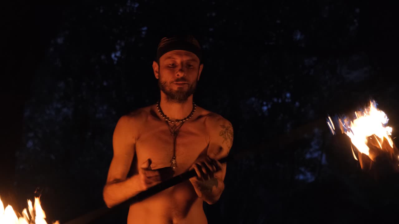 plano medio de un artista de fuego masculino haciendo malabarismos con palos de fuego, también conocidos como dragonstaff, que lo iluminan sobre un fondo oscuro
