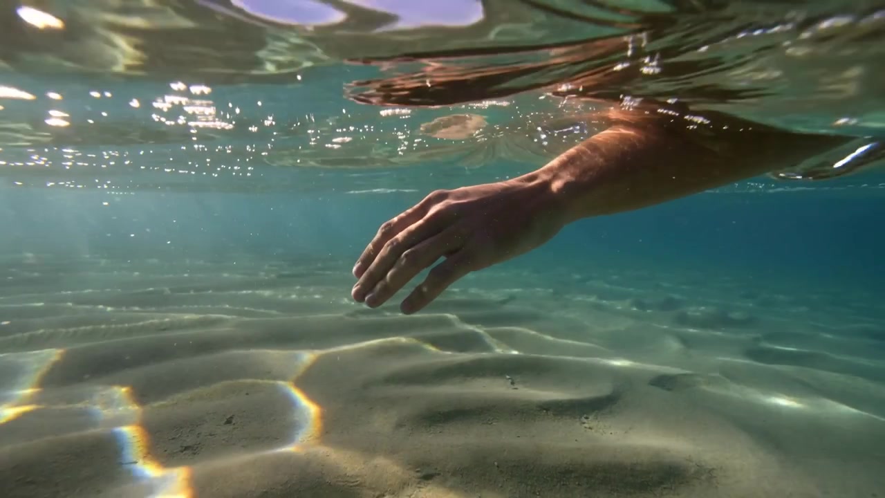 Entrando dalla superficie, un avambraccio nudo scivola sotto il mare poco profondo mentre percepisce le increspature della sabbia.