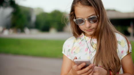 Niña-Sonriente-Jugando-Juegos-En-El-Teléfono-Móvil-Al-Aire-Libre.-Chica-De-Pie-En-El-Parque