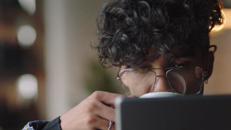 close up attractive young mixed race man student using tablet computer in cafe browsing online reading social media messages enjoying drinking coffee relaxing wearing stylish glasses