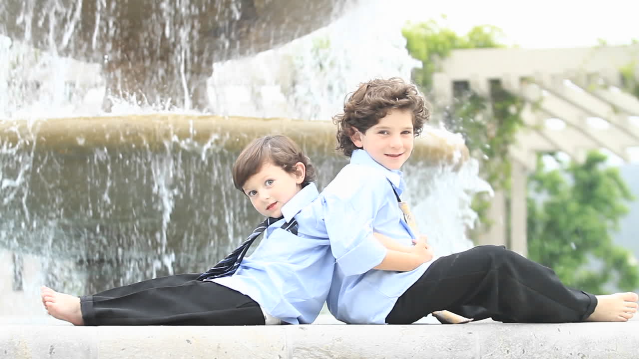 пара мальчиков в формальной одежде сидят спина к спине перед фонтаном