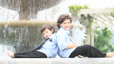 a pair of boys in formal dress sit back to back in front of a fountain