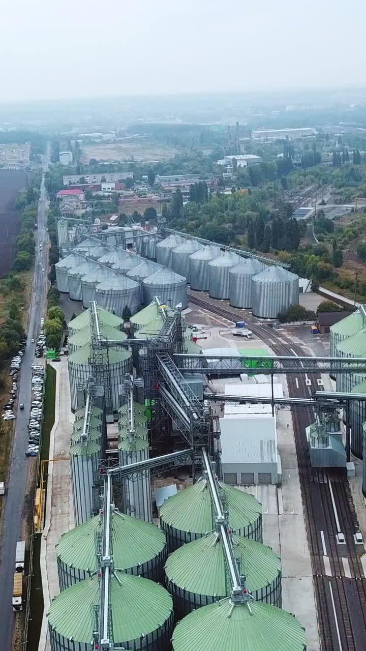 The territory of a huge plant divided by railways. Silo tanks of granary elevator at the backdrop of farmlands and countryside. Vertical video