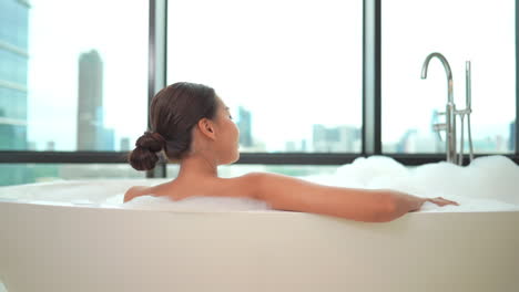 a woman sits in a luxurious bubble bath while looking out at a modern city skyline