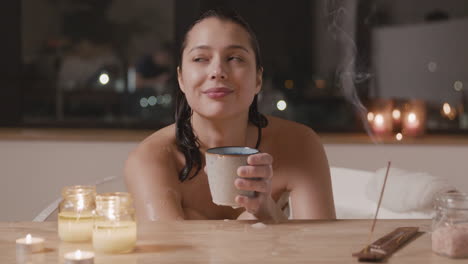 close up view of a woman taking a bath, while drinking from a cup at a table decorated with candles and incense 1