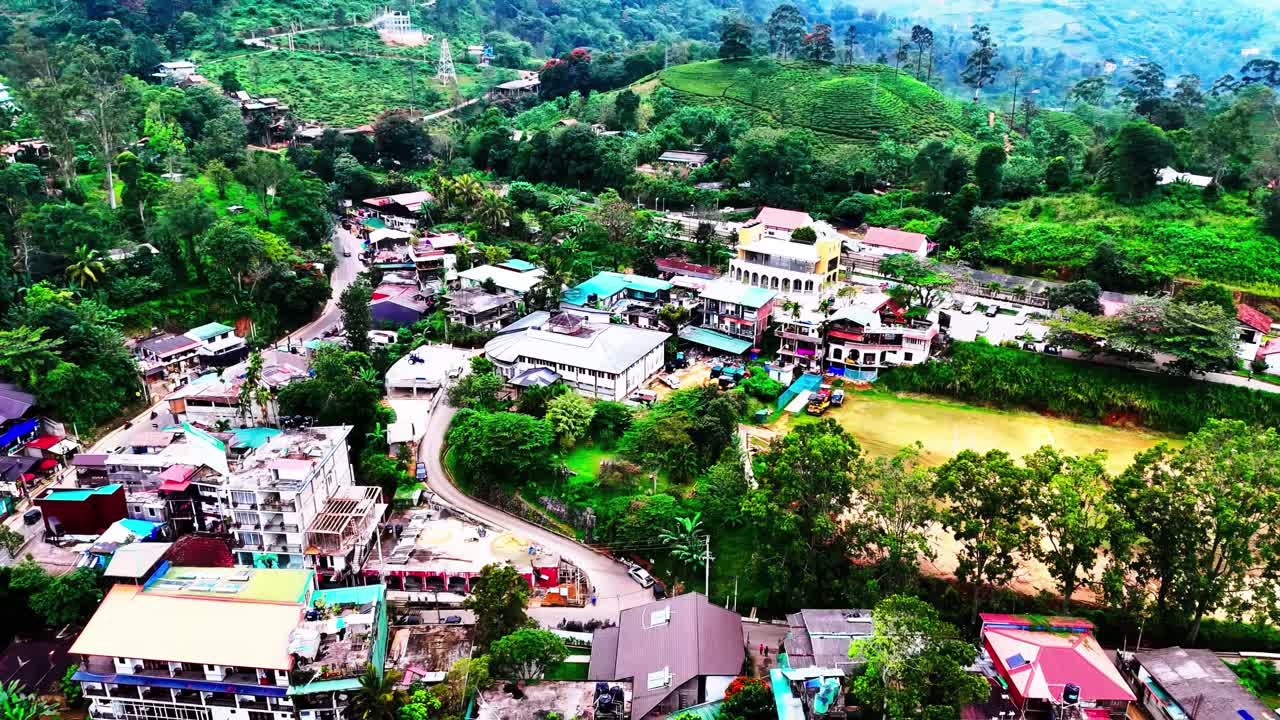 A vivid aerial captures a Sri Lankan village dotted with colorful buildings, a central sports field, winding roads, and lush surrounding hills blending human settlement and nature.